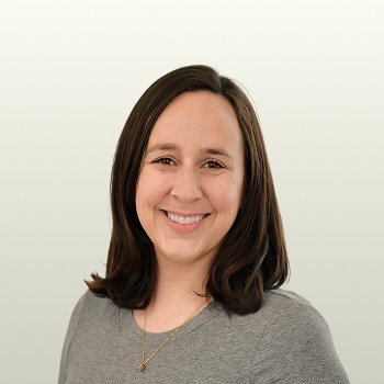 A woman with straight, shoulder-length brown hair smiles at the camera. She is wearing a gray top and a gold necklace, standing against a plain, light-colored background.