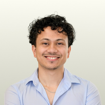 A person with curly dark hair, a short mustache and beard, wearing a light blue collared shirt and a chain necklace, smiling at the camera against a plain light background.