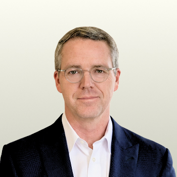 A man with short gray hair and glasses, wearing a dark suit jacket over a white collared shirt, looks directly at the camera against a plain light background.