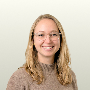 A woman with long blonde hair, wearing clear eyeglasses and a brown sweater, smiles at the camera against a plain, light background.