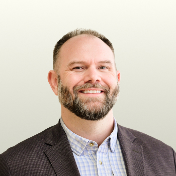 Smiling man with a beard wearing a blazer over a checkered shirt, posing in front of a plain, light background.