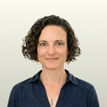 A woman with short, curly dark hair is wearing a navy blue shirt and a necklace, facing the camera with a neutral background.