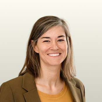 A woman with straight, shoulder-length brown hair and light highlights smiles. She is wearing a brown blazer over a mustard yellow top, with a light gray background behind her.