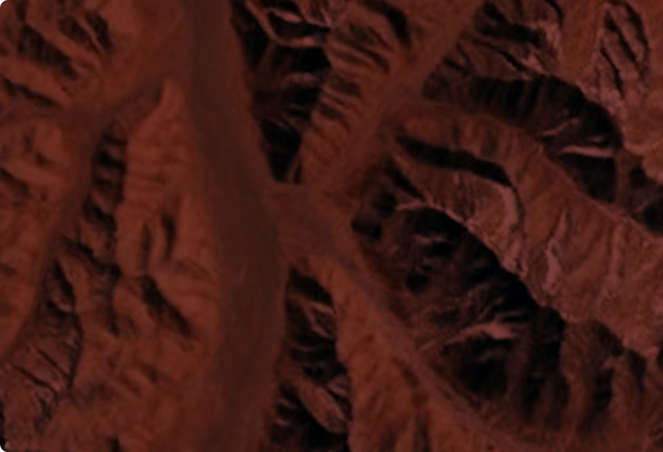 Aerial view of rugged, reddish-brown terrain with deep valleys and sharp ridges, resembling a rocky or canyon landscape. Shadows highlight the uneven, jagged surface.