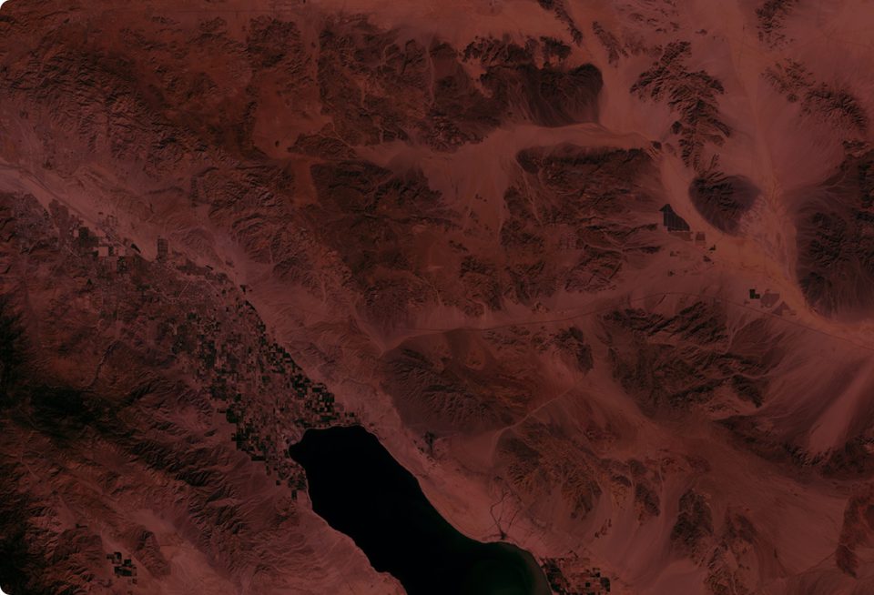 Satellite image showing a dark body of water surrounded by mountainous desert terrain, with a patchwork of green and brown agricultural fields at the edge of the water, under reddish lighting.