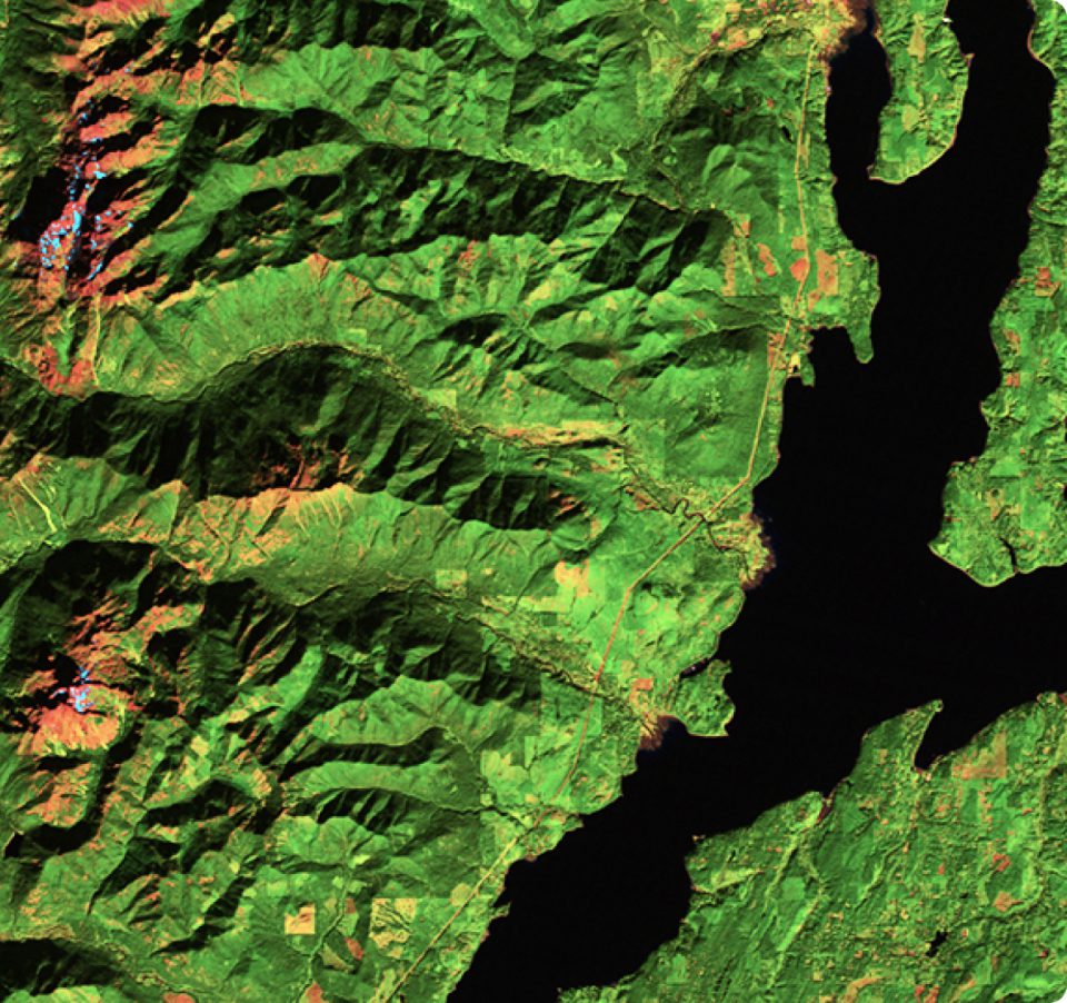 Satellite image showing a green, mountainous landscape with ridges and valleys, intersected by a large dark blue river or lake. Sparse small settlements and lighter patches of vegetation are visible.