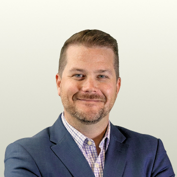 A man with short light brown hair and a trimmed beard, wearing a blue blazer over a checked shirt, smiling with arms crossed in front of a plain light gray background.