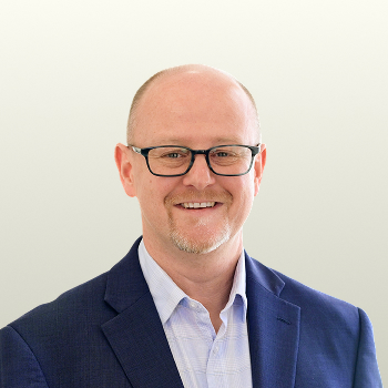 A middle-aged man with light skin, glasses, and a short beard smiles at the camera. He is wearing a blue blazer and a light-colored shirt, standing against a plain light background.