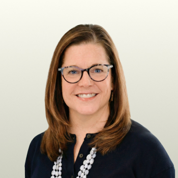 A woman with straight brown hair and glasses smiles at the camera. She is wearing a navy blue top and a white beaded necklace, with a light, plain background.