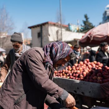 A person wearing a scarf and jacket bends over a cart filled with apples at an outdoor market, with other people and produce visible in the background under a clear blue sky.