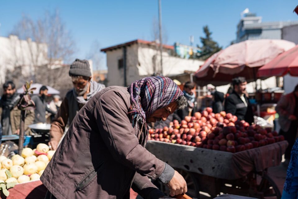 A person wearing a scarf and jacket bends over a cart filled with apples at an outdoor market, with other people and produce visible in the background under a clear blue sky.