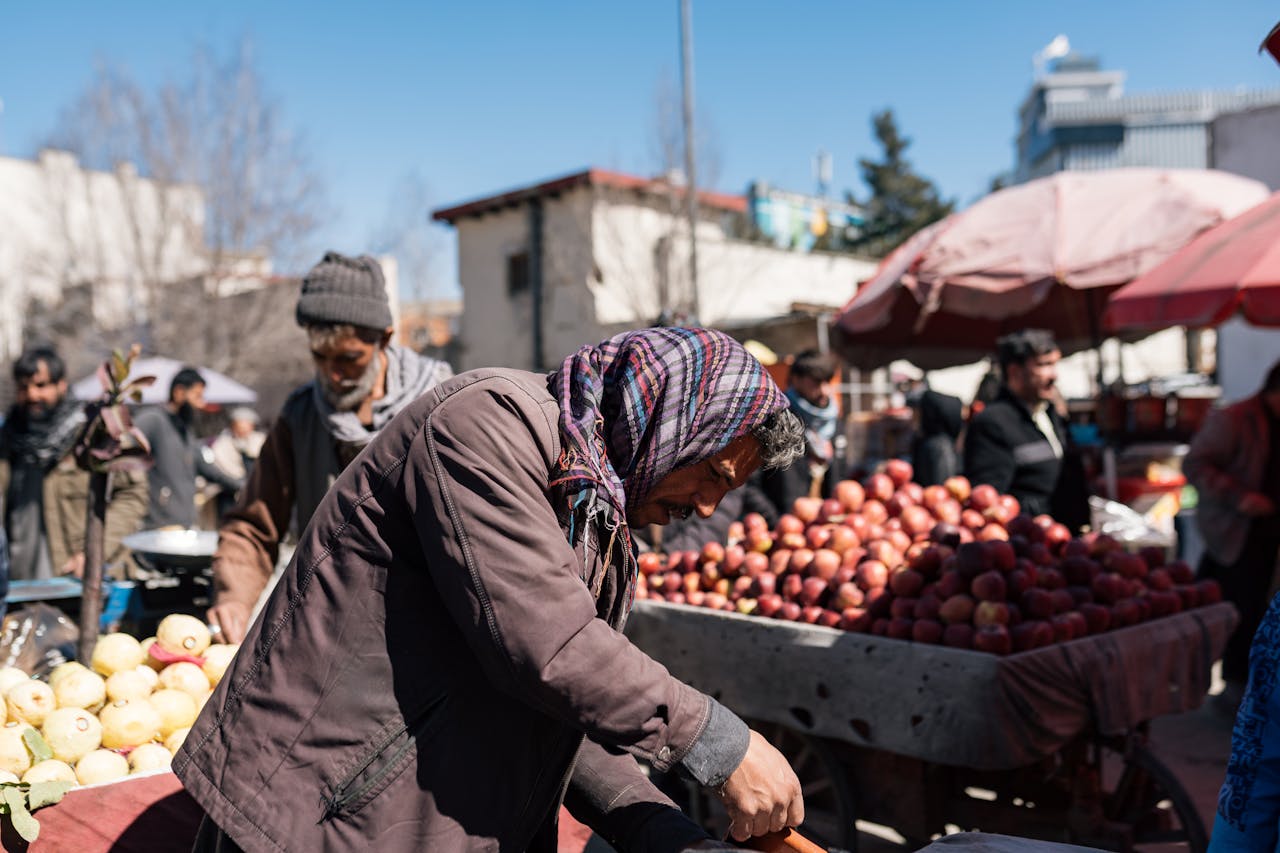 A person wearing a scarf and jacket bends over a cart filled with apples at an outdoor market, with other people and produce visible in the background under a clear blue sky.