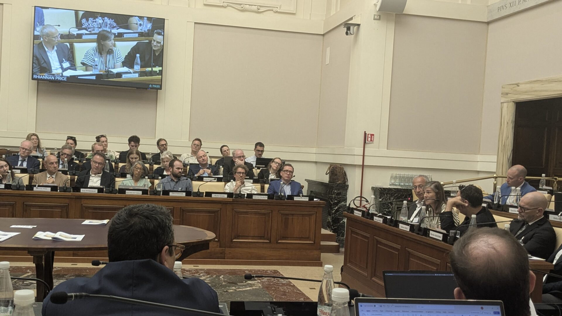 A conference room filled with people listening to a speaker. Attendees sit at tables and face a panel, with a monitor displaying a close-up of the discussion. The setting appears formal and academic.