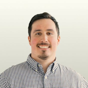 Man with short dark hair and a trimmed goatee smiling, wearing a checked button-up shirt, standing in front of a plain light background.